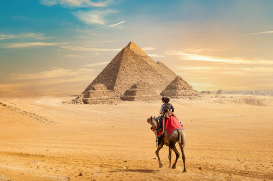 A person rides a camel in the Egyptian desert with the Great Pyramid in the background